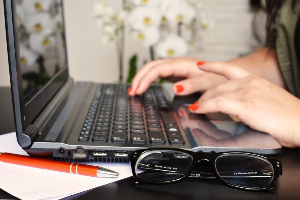 unas manos con uñas pintadas de rojo teclean en una laptop. al lado hay unas gafas de leer y una pluma fuente.
