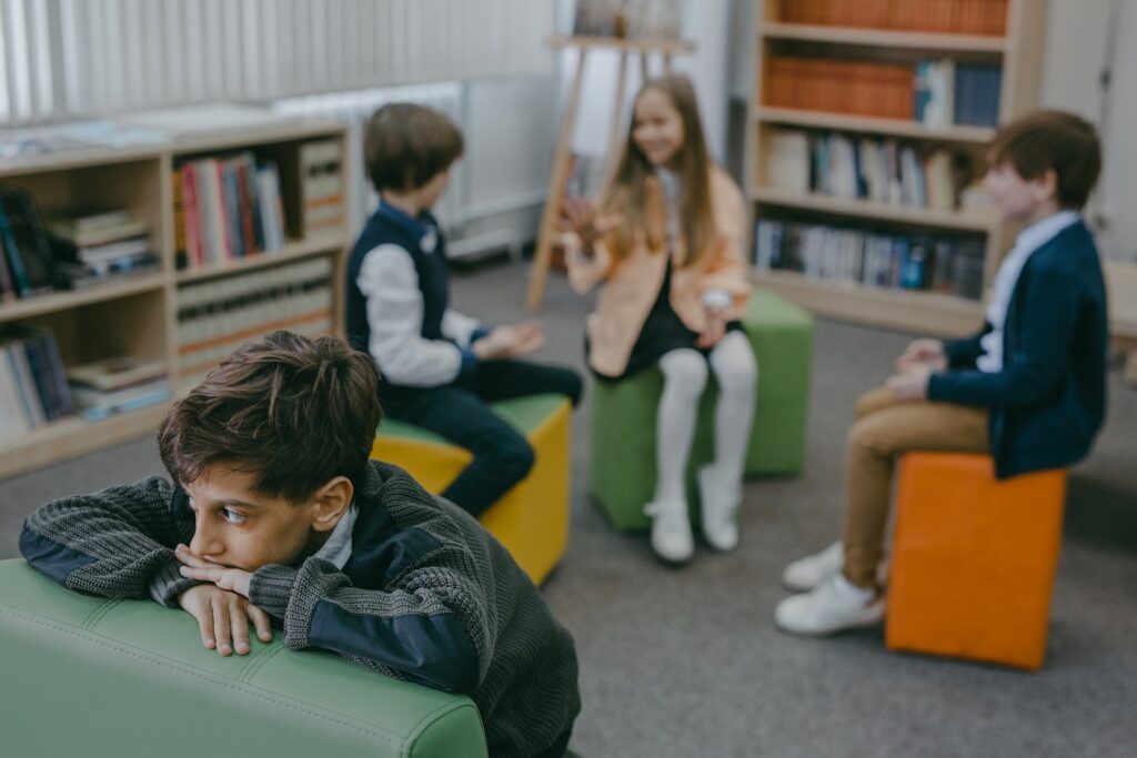 Niño triste se aísla de sus compañeros en el aula, mientras ellos conversan entre sí.