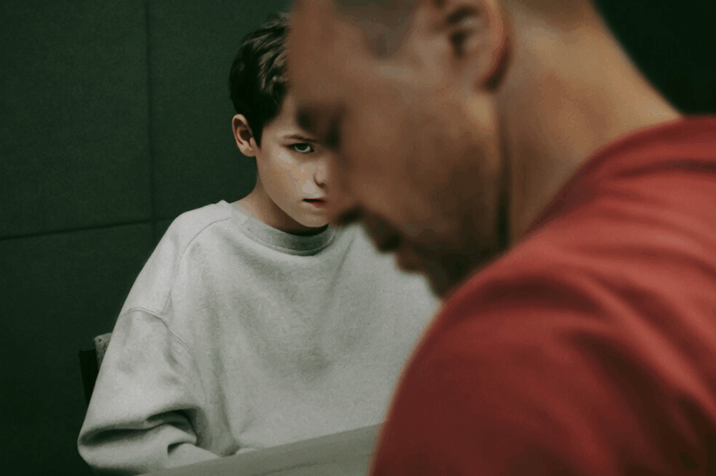Imagen de la serie Adolescencia de Netflix en donde un joven ve hacia la camara maliciosamente mientras también vemos el perfil de un adulto.