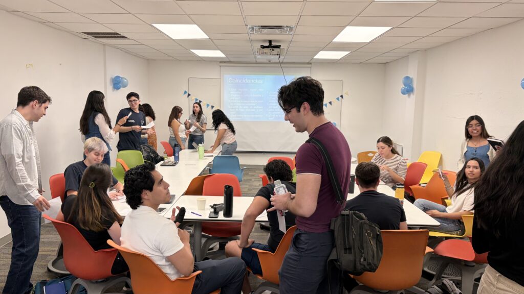 Estudiantes universitarios participan en una actividad colaborativa en el aula, trabajando en equipos y conversando alrededor de mesas.